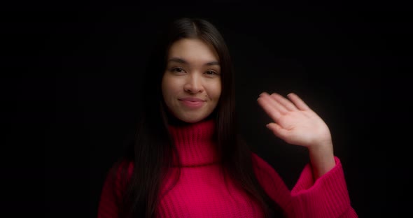 Smiling Asian Woman in a Bright Pink Sweater Waves Her Hand