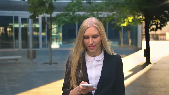 Cheerful Business Woman with Phone Outdoors. Content Blond Lass in Black Jacket Holding Smartphone