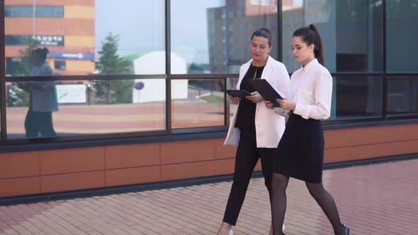 Two Young Business Girls Walking Down the Street and Discussing a Working Project