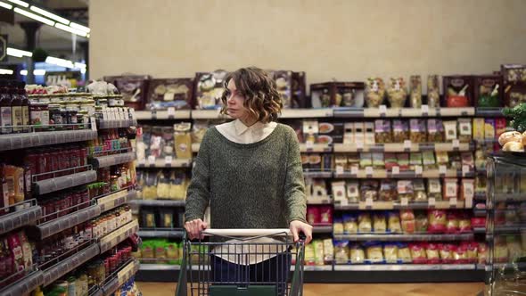 Portrait of a Woman with Wavy Hair Wearing Casual is Driving Shopping Trolley Through the Row