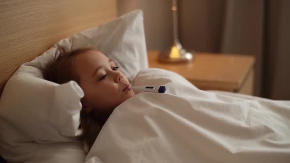 Closeup Face of Little Sick Girl with High Temperature Lying in Bed with Thermometer in Her Mouth