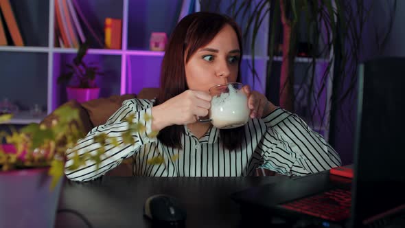 Young Woman Drinks Coffee Sitting in Armchair Before Computer in Office