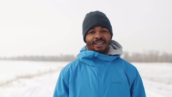 Black man in warm winter clothes stands outdoors at winter and looks at camera and smiles, front vie