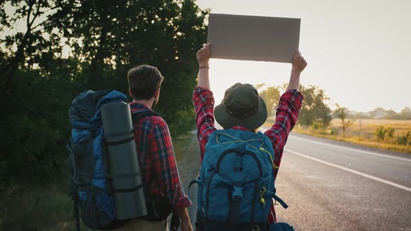 Active Tourists Hitchhiking on Empty Road Standing with Backpacks and Signboard