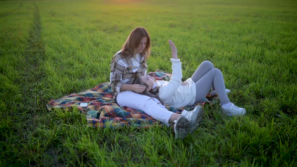 Loving tender mum with a cute daughter sitting on a blanket on a field at sunset