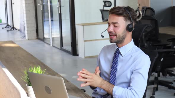 Cheerful young support phone male operator in headset, at workplace while using laptop