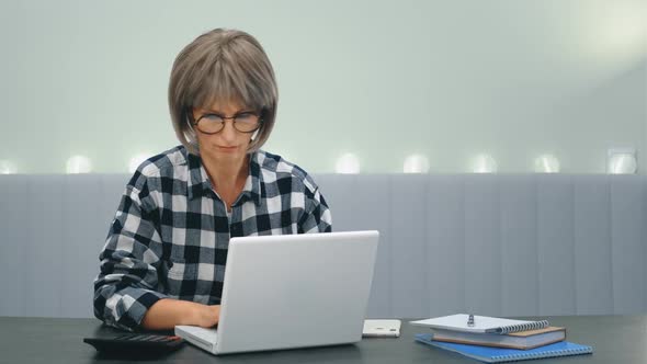 An Elderly Tired Woman with Glasses is Sitting at a Laptop and with Difficulty Overcoming Drowsiness