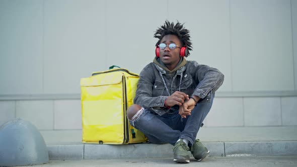 Carefree Young African American Man Sitting on Urban City Street with Yellow Insulated Food Delivery