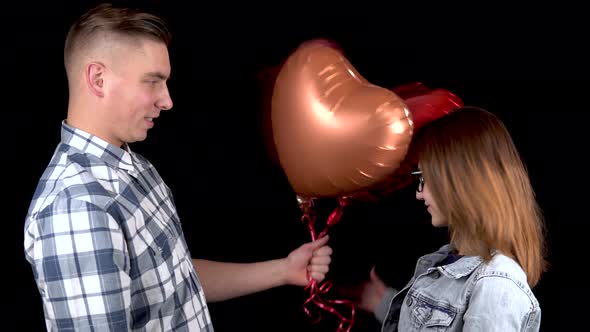 A Young Man Gives a Young Woman Heart-shaped Balloons. Man and Woman Kissed on a Black Background
