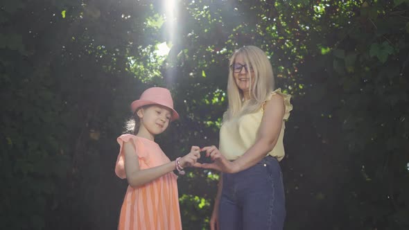 Cute Mature Blond Mother and Her Pretty Daughter Making Heart Shape with Their Fingers in the Summer