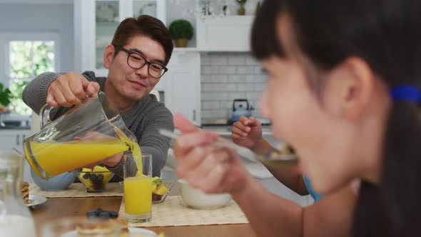 Happy asian father in kitchen having breakfast pouring juice for smiling son and daughter