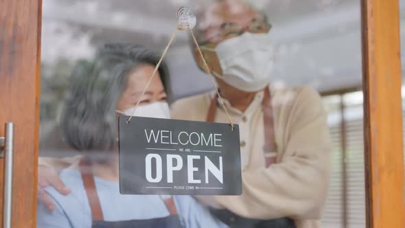 Asian Senior Couple wearing mask turning Welcome we are open sign on glass door to reopen business.