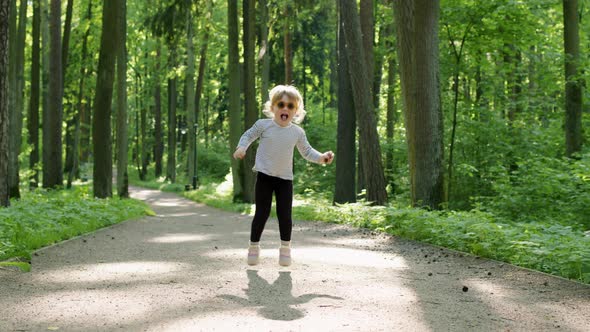 Cheerful Child in Striped Tshirt Jump on Ground on Summer Sunny Day in Park Among Tall Green Trees