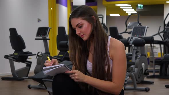 A Young Beautiful Woman Writes Into a Notebook in a Gym - Closeup