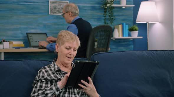 Senior Woman Reading Emails on Digital Tablet Sitting on Sofa at Home