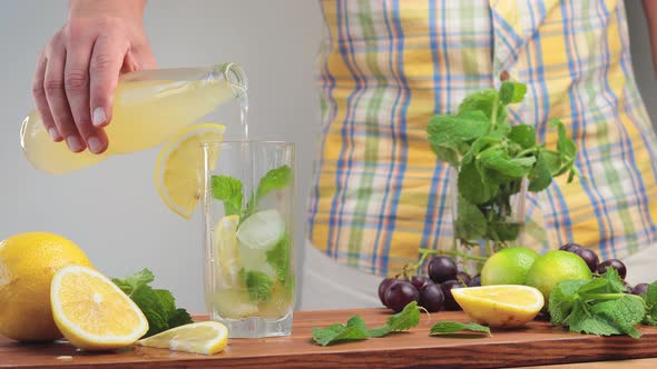 Pouring lemonade from bottle into glass with mint, ice cubes and lemon slices, close up view