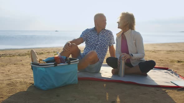 Happy Senior Couple Having Picnic on Sea Beach Drinking Coffee or Tea in Thermos Bottle