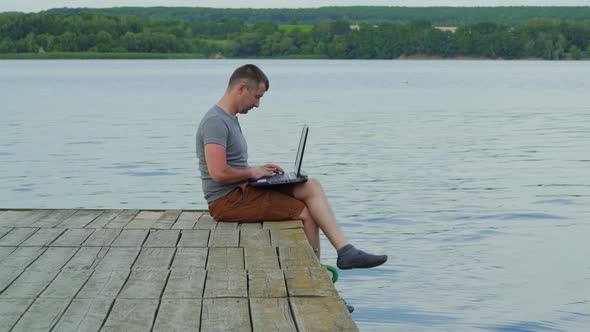 Young Man Working on Laptop While Sitting оn Old Pier