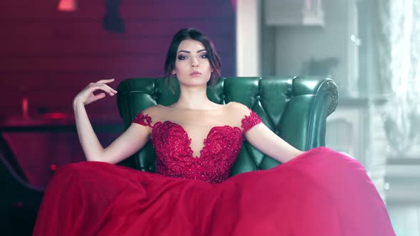 Portrait of Sexy Fashion Woman in Red Evening Glamour Dress Sitting on Armchair Looking at Camera