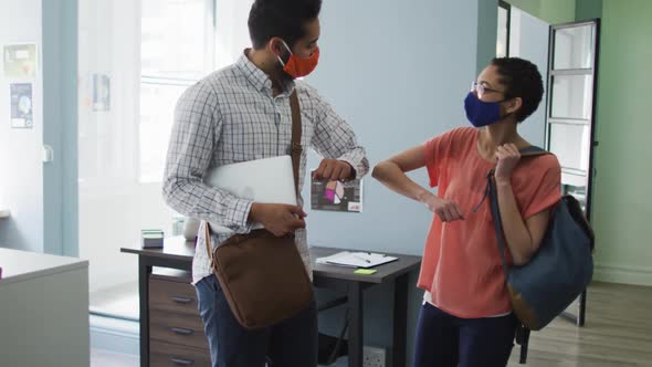Diverse male and female colleague wearing face masks greeting each other by touching elbows at moder