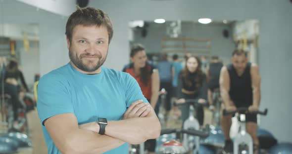 Happy Confident Coach Posing at the Background of Blurred Sportive People Riding Exercise Bikes in alt