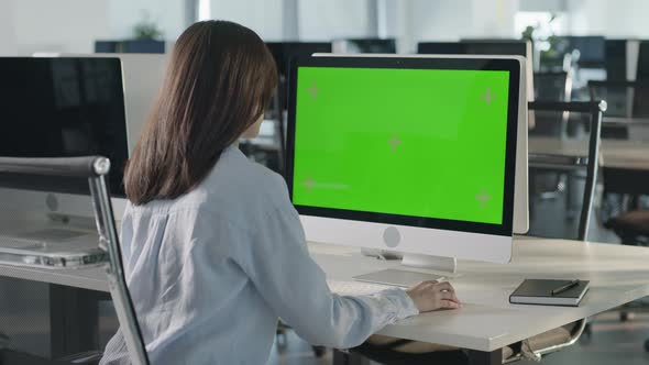 Asian Business Woman Typing on Desktop Computer with Green Mockup Screen While Sitting at His Desk