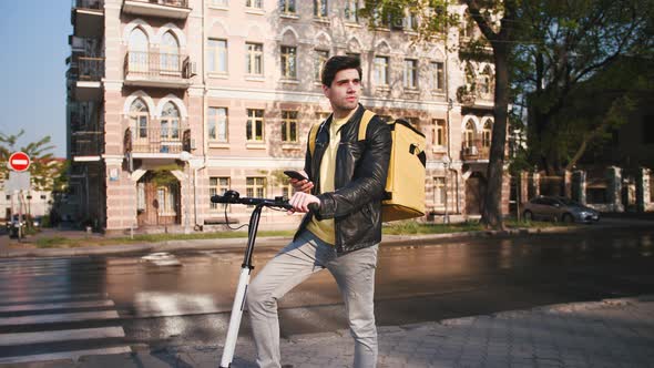 Delivery Man with Yellow Bagpack Standing with Electric Scooter in City and Using Smartphone to Find