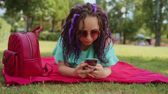 Young woman lying on a blanket blowing uses a smartphone in the park.