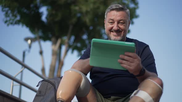 Joyful Bearded Male Amputee Smiling Surfing Social Media on Tablet Sitting Outdoors