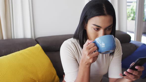 Gender fluid male drinking coffee and using smartphone while sitting on the couch at home