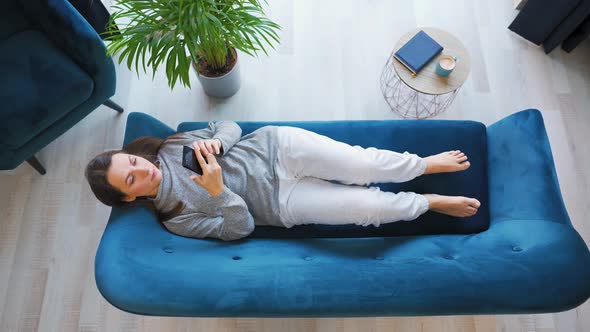 Overhead Shot of Woman Goes to the Sofa and Lies Down on It Holding Smart Phone Using Mobile Apps