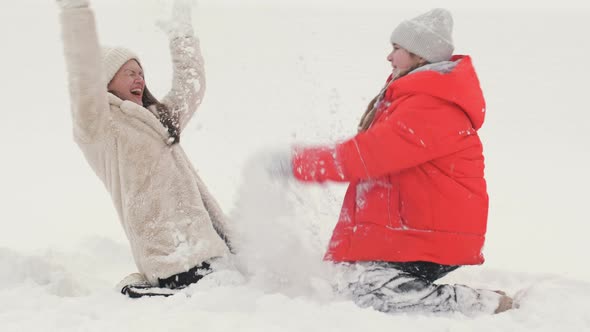 Young Woman with Her Teenage Daughter Is Kneeling in the Snow and Cheerfully Sprinkling Snow on Each