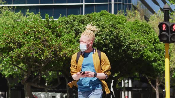 Albino african american man with face mask and dreadlocks walking and using smartphone