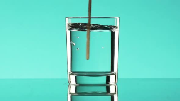 Clear Water Mixing Up with Stirring Rod in Glass Standing on Turquoise Background in Slowmotion