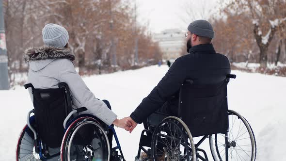 Disabled husband and wife sitting in wheelchairs holding hands resting in winter park, back view.