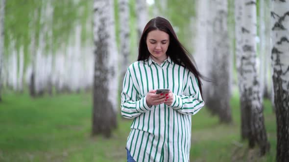 Beautiful Girl with a Smartphone in Her Hands Walks Along a Birch Grove in Spring