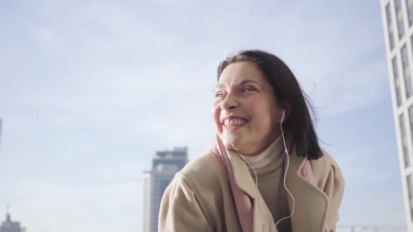 Close-up of Young Caucasian Woman in Headphones Standing on City Street and Smiling. Smiling