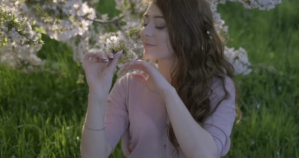 Girl Dressed in Airy Pink Dress Smelling the Flowering Branch of Apple Tree and Smiling