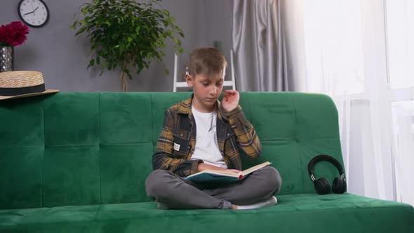 Teen Boy is Tired to Read Uninteresting Book, Sitting on Green Soft Couch at Home