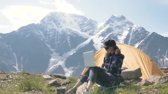 Lady Sits on Hill Top and Types on Laptop Against Mountains