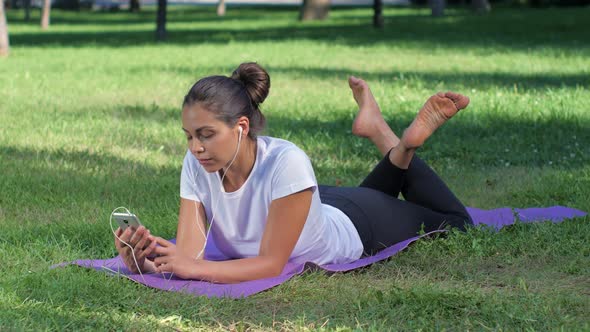 Girl with Headphones and with the Phone in Her Hand Lying on the Yoga Mat