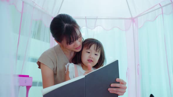 Asian mom read storytale to young baby girl daughter in tent at home.