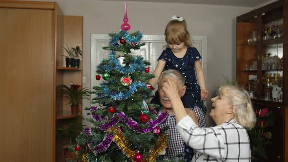 Kid Girl with Senior Grandmother and Grandfather Decorating Artificial Christmas Tree with Toys