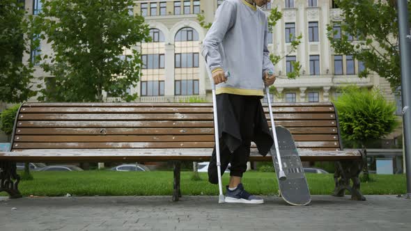 Attractive Man Sits on the Bench in the Park Putting His Crutches and Skateboard Nearby. Active Life