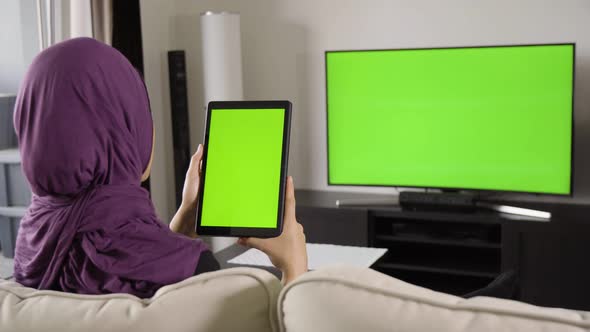 A Muslim Woman Looks at a Tablet (Vertical) and TV (Both Green Screen) As She Sits on a Sofa