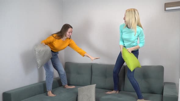 Two Friends or Sisters Fight with Pillows Jumping on the Couch at Home