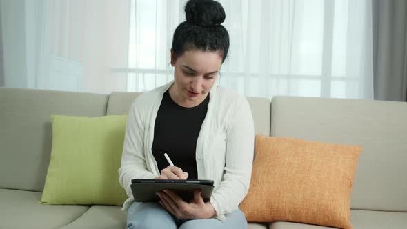 Woman Sits on the Sofa at Home Drawswrites and Uses a Pen with a Digital Tablet Computer