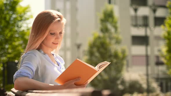 Young Beautiful Blonde Girl in Summer Day is Reading a Book Sit on a Bech at Urban City Park