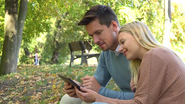 A Young Attractive Couple Works on a Tablet and Talk in a Park on a Sunny Day - Closeup