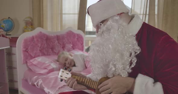 Santa Claus in Red Costume with False Beard Playing Ukulele As Little Blond Caucasian Girl Sleeping